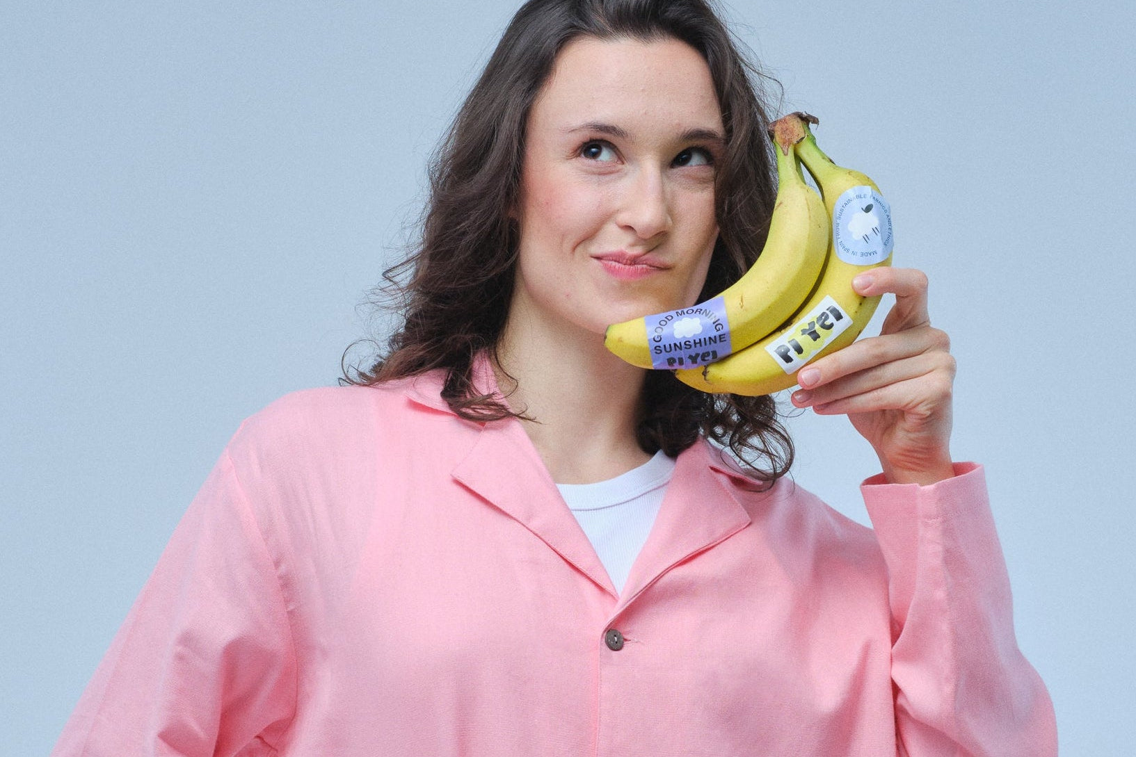 Woman in pink outfit holding a bunch of bananas with a phone on a light blue background